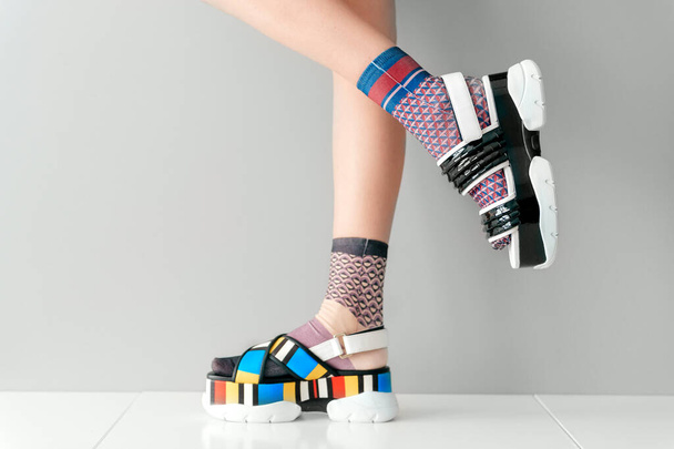 Beautiful female legs in mismatched trendy socks standing in two different fashionable high wedge leather sandals on white surface. Odd disargonized  young girl wearing high sole summer stylish shoes - 写真・画像