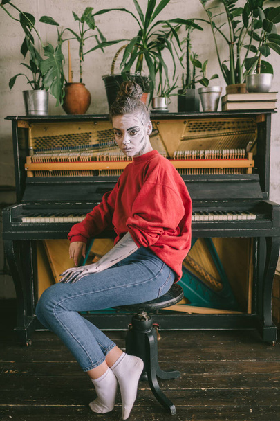 Srrange young girl with drawings on her face sitting on chair in interior room with piano on background. - Фото, зображення