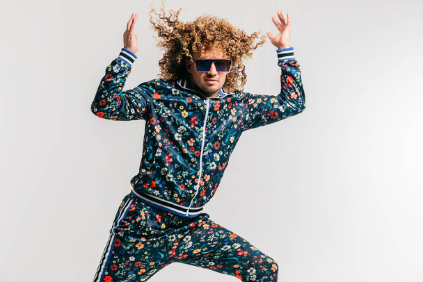 Excited adult funny man in stylish vintage clothes posing on white studio background. 80s fashion. Funky guy in tracksuit and sunglasses expressive indoor unusual portrait. Shouting cheerful male. - 写真・画像