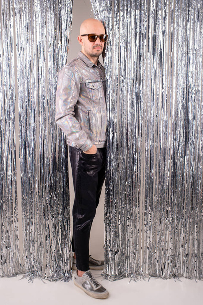 Beautiful caucasian man without hair in a glittering white shirt, black leather pants, grey shoes and black sunglasses shows different poses for a fashion magazine on tinsel background. - Φωτογραφία, εικόνα
