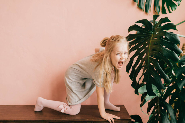 Beautiful little blonde long haired child girl in white tights posing like wild tiger with excited funny face on pink wall background.  Exalted female child shows screaming wild cat in the jungle - Photo, Image
