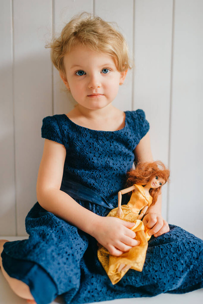 Trubschachen, Switzerland - December 16, 2019: Pretty caucasian little girl with short fair hair in blue dress holds her lovely barbie toy, sits in bright baby room and smiles - Foto, imagen