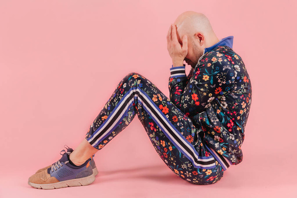 Adult unhappy sad bald depressed man in fashionable trendy tracksuit isolated on pink background.  Conceptual psychological portrait of stylish despaired boy siiting on ground indoor. Odd crying boy - Zdjęcie, obraz