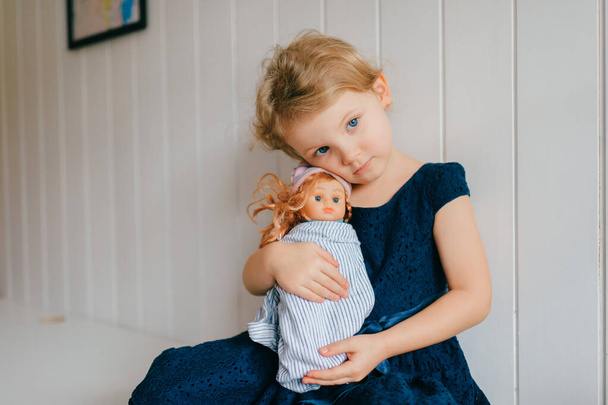 Trubschachen, Switzerland - December 16, 2019: Pretty caucasian little girl with short fair hair in blue dress holds her lovely barbie toy, sits in bright baby room and smiles - Foto, imagen