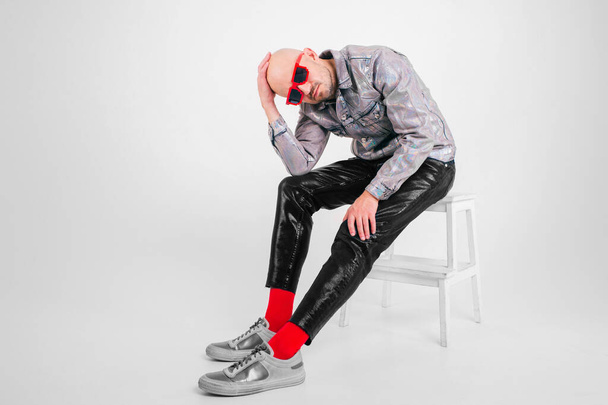 Beautiful caucasian man without hair in grey shirt, black leather trousers, black sunglasses, red socks and grey trainers poses for the camera isolated on white background. - Photo, image