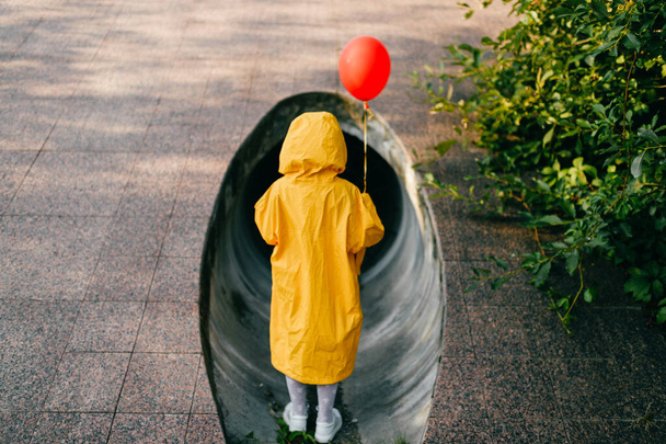 Portrait of pretty litle girl in big adult oversized yellow raincoat with red balloon in hand standing and sitting against sewage tunnel pipe. Horror movie concept. Funny child posing in summer day - Zdjęcie, obraz