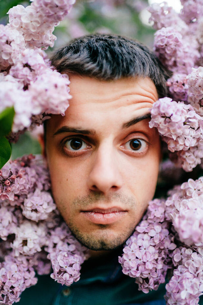 Closeup odd unusual male portrait outdoor at nature in beautiful lilac bushes.  Adult funny man face grimace surrounded by blooming flowers. Emotions and facial expxression. Clowning and fooling - Foto, Imagem