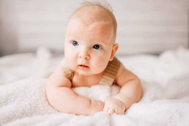 Adorable lovely newborn male baby boy with smiling emotional  happy face lifestyle indoor portrait. Funny infant child lying on stomach on bed with white wall on background.  Carefree childhood - Φωτογραφία, εικόνα
