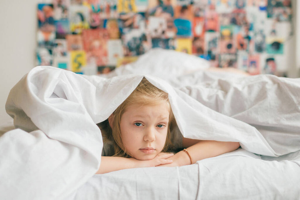Young adorable blonde girl with funny face lying on bed under white blanket and look at camera with sad eyes. Little female child with pensive face indoor lifestyle portrait. Little  girl portrait - Fotografie, Obrázek
