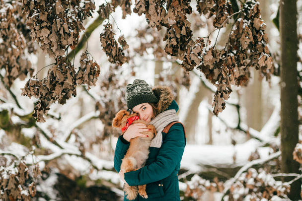 Young positive girl in winter clothes holding in hands her adorable pomeranian puppy in snowy forest - Φωτογραφία, εικόνα