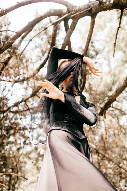 Young beautiful elegant girl in black vintage dress with veil posing among trees outdoor. - 写真・画像