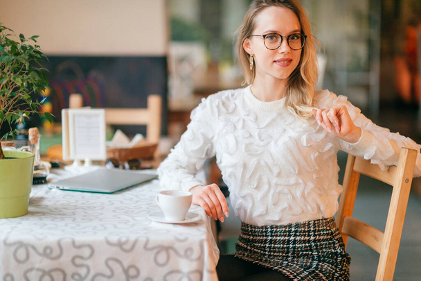 Young beautiful blonde lady in white sweater and glasses sitting in caffe and posing for camera with positive face expression. - Foto, afbeelding