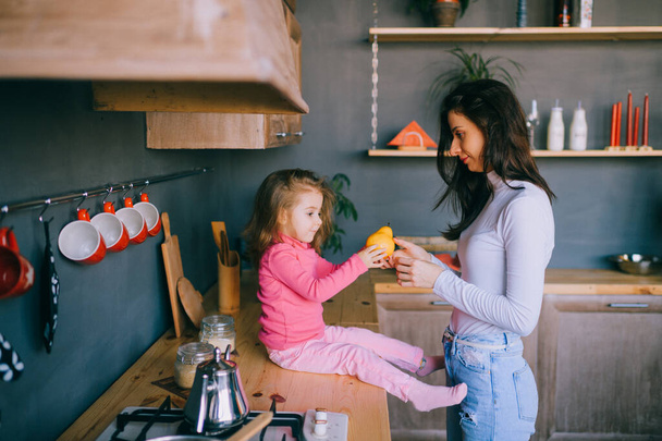 Adorable young woman playing with her little funny daughter in kitchen. Portrait of pretty mother hugging, carrying and watching her litle female child. Happy family indoor lifestyle portrait. - Foto, Bild