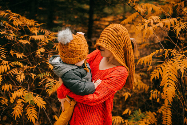 Handsome caucasian mother in warm grey jacket, black trousers and grey trainers hugs and rejoices with her little son in warm bright clothes. - Photo, image