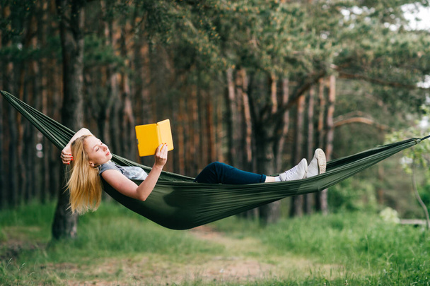 Young beautiful blonde girl in hammock reading e-book. Pretty woman leisure lifestyle portrait at nature ountdoor. Female relax in forest. Adorable teen expressive face. Tourism in summer camp. Hobby - Photo, Image