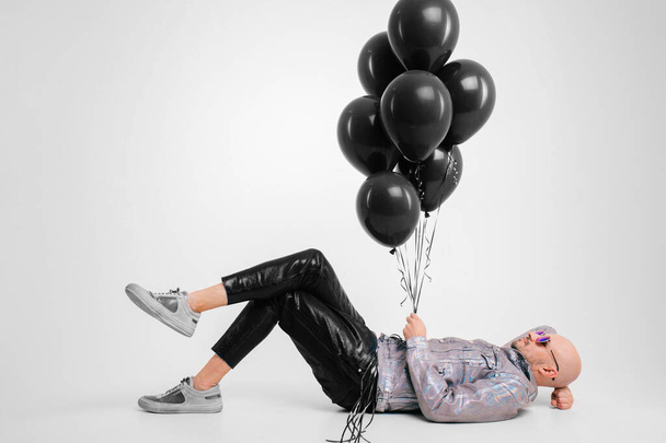 Beautiful caucasian man without hair in grey shirt, black leather trousers and grey trainers with black sunglasses lies on the floor and dream isolated on white background. - Fotó, kép