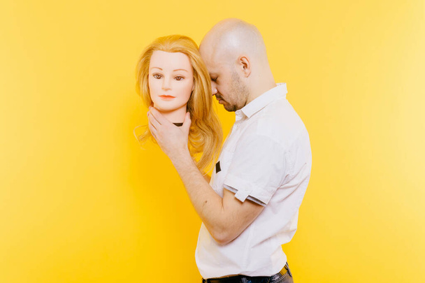 Excited odd bizarre strange unusual sad adult bald hairless man holding plastic blonde woman head. Funny male with girlfriend isolated on yellow. Baldness after chemotherapy. Cancer hair loss concept - Фото, изображение