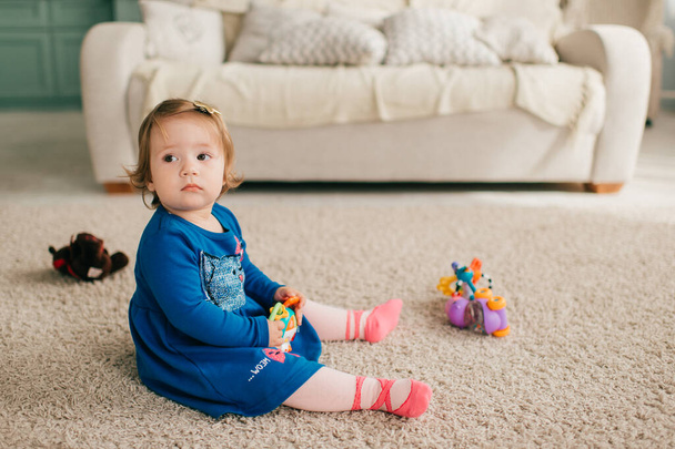 Malá roztomilá běloška v atraktivních šatech sedí na podlaze a koberci, hraje si s různými hračkami a úsměvy - Fotografie, Obrázek