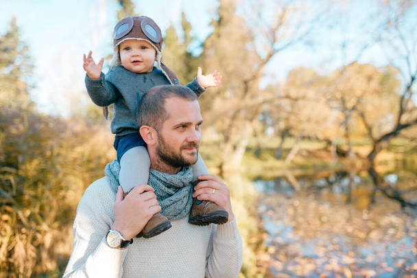 Happy father holding his beautiful son in pilot hat on his shoulders in autumn park - Фото, изображение