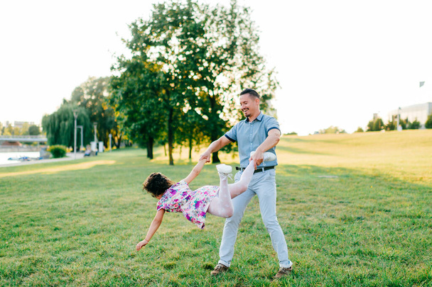 Father holding his daaughter hand with leg and whirling around. Excited family have fun in summer park. Dad playing with little female child on green meadow. Carefree happy childhood. Fatherhood  - Foto, imagen