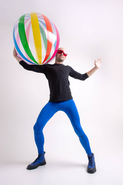 Fitness young man in blue sportive tights, black athletic shirt and stylish boots holding beach ball on his houlder against white studio background - Foto, imagen