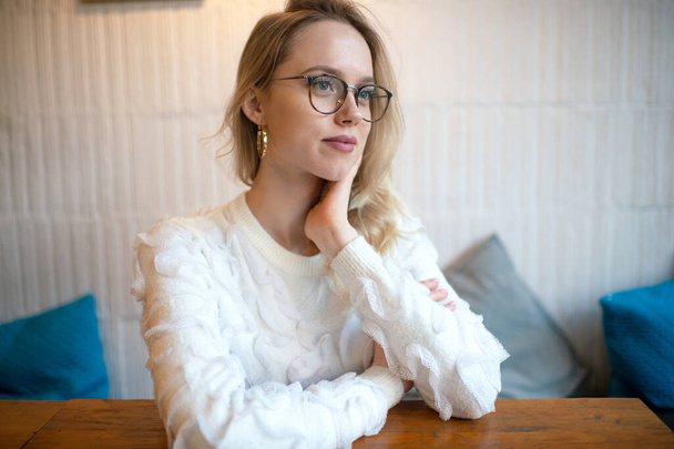 Indoor lifestyle portrait of cheerful charismatic caucasian girl in glasses wering white sweater. - Valokuva, kuva