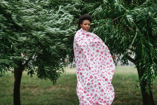 Beautiful dark skinned girl wrapped in sheet with fashion print posing at park with trees on background. - Φωτογραφία, εικόνα