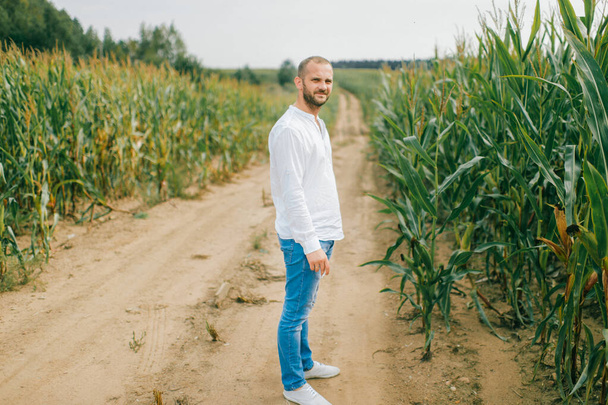 Beautiful young caucasian man in white shirt, blue jeans and white boots goes for a walk in the cornfield. - 写真・画像