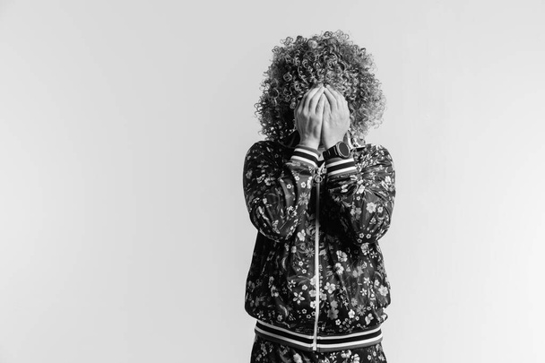 Unrecognizable man in fashionable modern tracksuit with flowers and afro hairstyle hidng  face with hands. Indoor portrait of unknown male depressed crying person. Isolated on white guy feeling shame - Fotó, kép