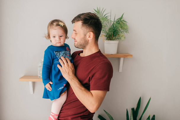 Caucasian handsome strong dad in a bright room hold his cute little daughter which sits on a shelf - Foto, imagen