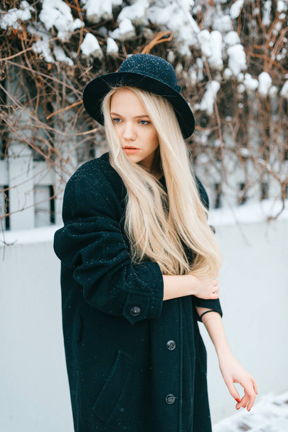 Stylish beautiful blonde girl in black clothes and hat posing near fence in the street - Photo, Image