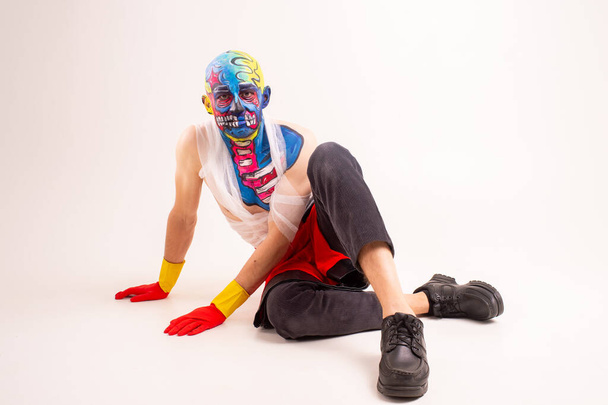 Strange man with makeup on his head and shoulder in white t-shirt, red and yellow gloves, red sits on a white floor - Foto, afbeelding