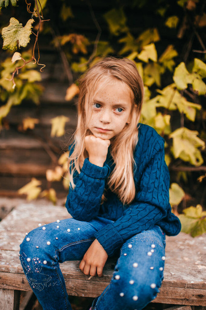 Caucasian handsome girl with long fair hair in blue hoody sits on an old chair in the garden near the tree in village and thinks about something - Foto, Imagem