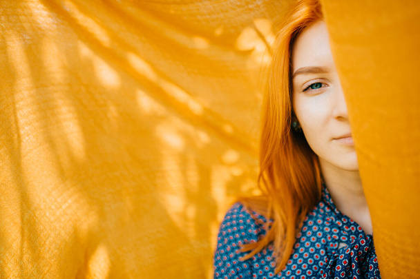Beautiful girl with red hair hiding half face behind orange blanket with abstract shadows on background. Mystery psychological woman portrait. Young pensive teen looking out of cover. Alter ego.  - Foto, Imagem