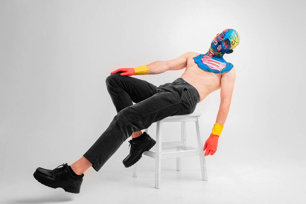 Portrait of a strange man with makeup on his head and shoulder in red-yellow gloves, black pants and boots, no T-shirt sits on a whitw chair - Foto, Bild