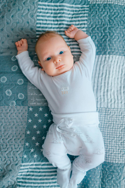 Adorable lovely newborn male baby with blue eyes lifestyle indoor portrait from above. Cute child with hands apart lying on back in pijama on bed at home. Little child with emotional face rest on sofa - Fotó, kép