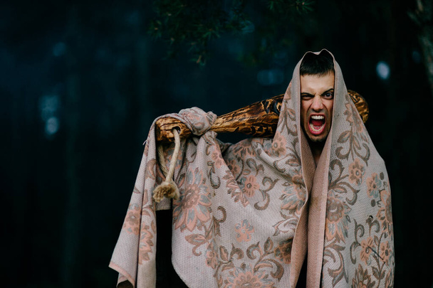 Funny young man with blanket on his head and big club on his shoulder fooling around in dark forest. Expressive excited boy face. Wild masculine strength. Cruel warrior - Foto, afbeelding