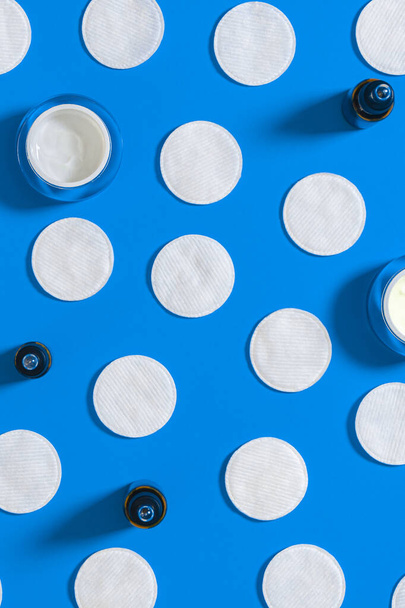 Hygiëneproducten: witte ronde wattenschijfjes en plastic sticks voor het reinigen van de oren liggen op een blauwe achtergrond naast gezichtscrème - Foto, afbeelding