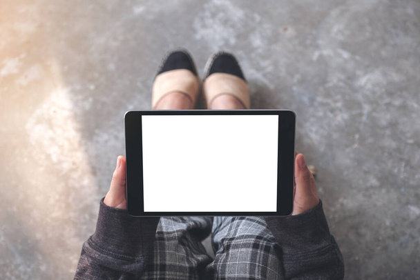 Mockup-Bild von oben von Frauenhänden, die einen schwarzen Tablet-PC mit leerem weißen Desktop-Bildschirm horizontal halten und verwenden, während sie auf dem Boden sitzen  - Foto, Bild