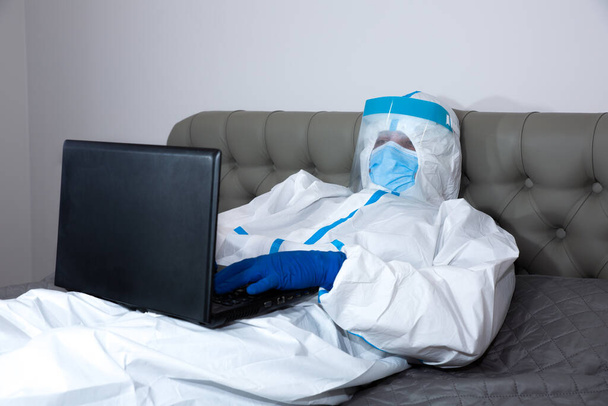 Doktor v lékařském ochranném obleku, brýlích, masce a rukavicích pracujících na notebooku, ležící doma na posteli. Ochrana proti epidemii viru. Koronavirus (COVID-19). Koncept zdravotní péče. - Fotografie, Obrázek