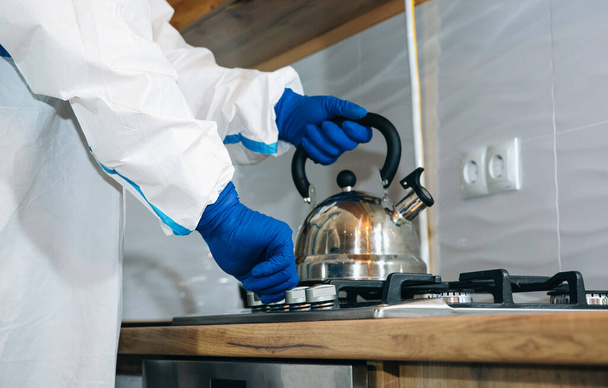 Doctor wearing medical protective suit, goggle, mask, and gloves preparing tea on the kitchen. Teapot on the gas stove. Protection mers by virus epidemic. Coronavirus (COVID-19). Healthcare concept. - Foto, afbeelding