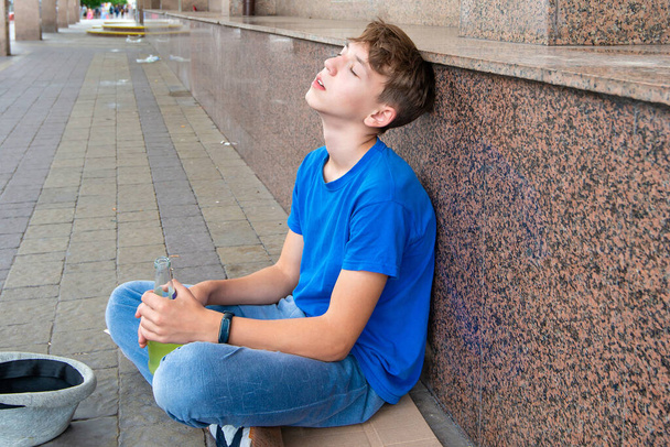 Juoppo teini-ikäinen kerjäläinen istuu drinkki kädessään ja kerjää rahaa. Teini-ikäisen alkoholismin käsite. - Valokuva, kuva