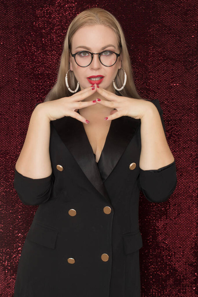 Beautiful young blonde woman in glasses in a black dress-jacket posing on a red shiny background. Hearts on her cheeks - Photo, Image