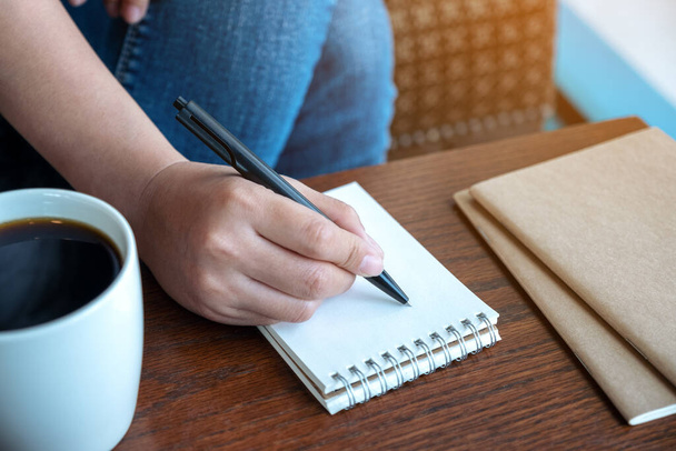 Immagine ravvicinata della mano di una donna che scrive su taccuino bianco con tazza di caffè su tavolo di legno - Foto, immagini