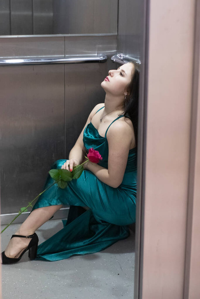 sexy confident brunette woman in elegant aquamarine colored dress with red rose in grey elevator. copy space - Fotoğraf, Görsel