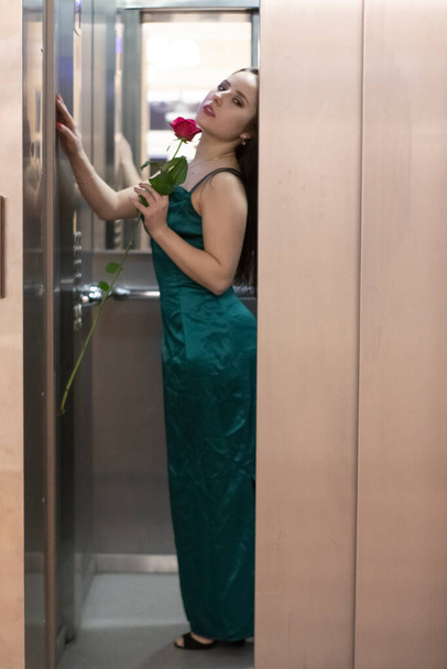 sexy confident brunette woman in elegant aquamarine colored dress with red rose in grey elevator. copy space - Foto, afbeelding