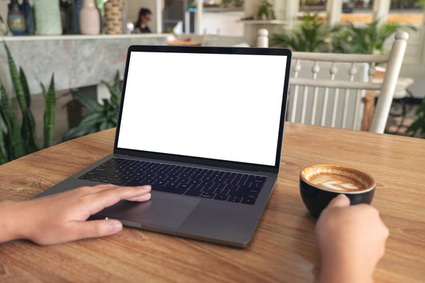 Imagen burlona de las manos de la mujer escribiendo y tocando el touchpad portátil con pantalla de escritorio blanco en blanco mientras bebe café en la mesa de madera en la cafetería - Foto, Imagen