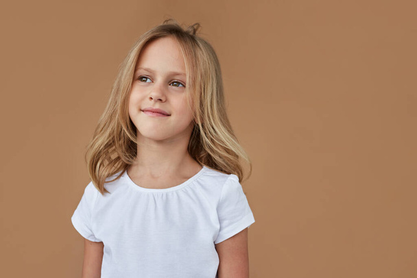 Closeup front portrait of pretty little girl with wavy blond hair, dressed in white clothes, over beige background. - Photo, Image