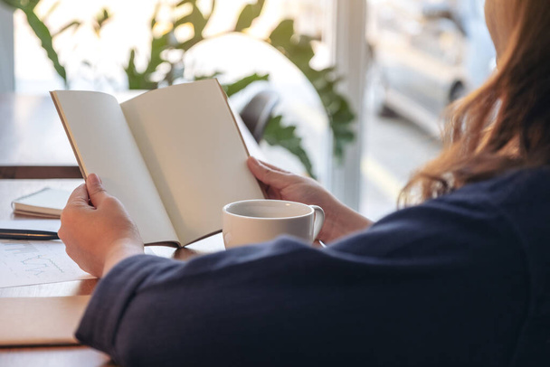 Immagine ravvicinata di una donna che tiene in mano e apre un quaderno bianco con tazza di caffè e carte sul tavolo - Foto, immagini