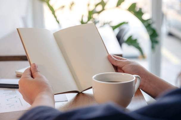 Imagem de perto de uma mulher segurando e abrindo um caderno em branco com xícara de café e papéis na mesa - Foto, Imagem
